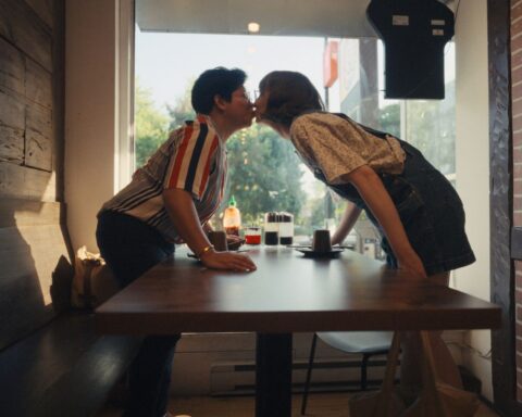 Two women--one Filipina, one white--stand up and lean over a table to share a kiss.