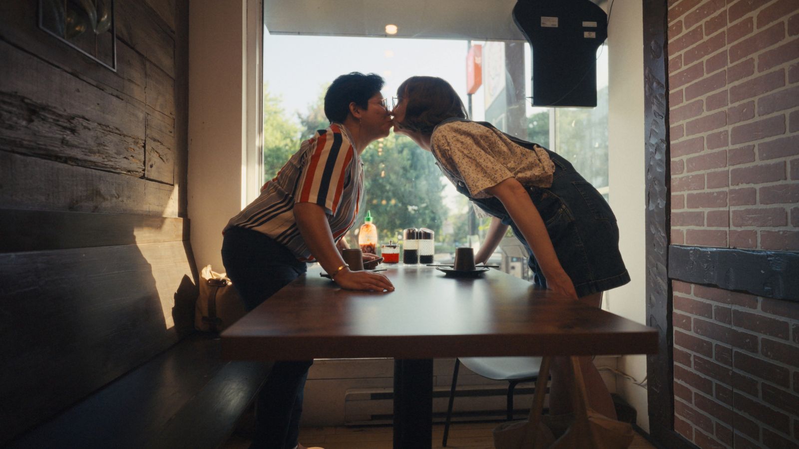 Two women--one Filipina, one white--stand up and lean over a table to share a kiss.