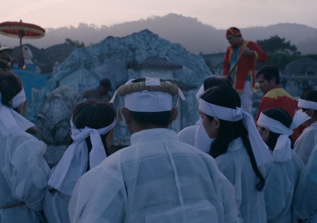 An image depicts a crowd of people during a Vietnamese mourning ritual. They are all wearing white robes and white headbands. The sun is beginning to set and the light is low.