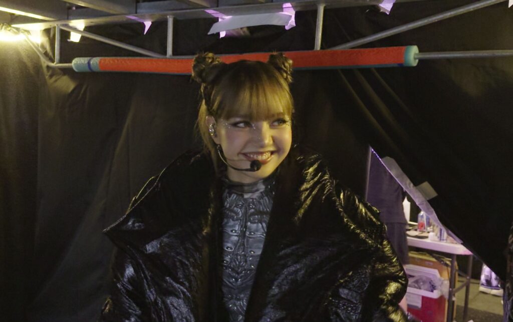 Thai musical artist Lisa appears backstage, ready to go on a show. She has a microphone headset and is smiling. She is wearing a large black puffy coat and silver bodysuit.