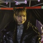 Thai musical artist Lisa appears backstage, ready to go on a show. She has a microphone headset and is smiling. She is wearing a large black puffy coat and silver bodysuit.