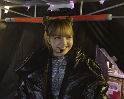 Thai musical artist Lisa appears backstage, ready to go on a show. She has a microphone headset and is smiling. She is wearing a large black puffy coat and silver bodysuit.