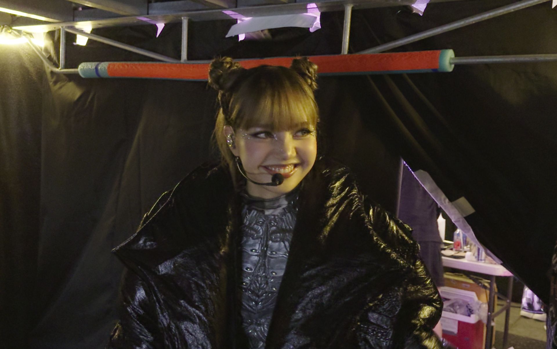 Thai musical artist Lisa appears backstage, ready to go on a show. She has a microphone headset and is smiling. She is wearing a large black puffy coat and silver bodysuit.