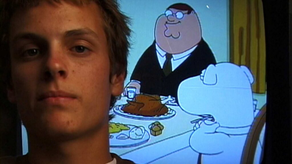 The face of a white man with an early 2000s' hairstyle is pictured beside a fuzzy TV screen showing a scene from Family Guy, where an overweight man in a suit sits at a table with a white dog.
