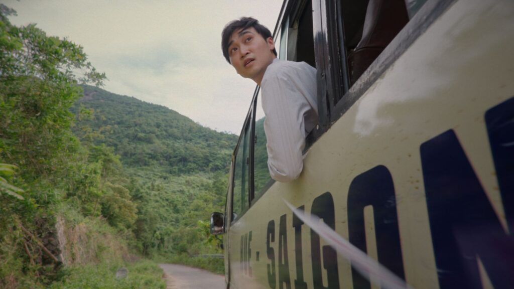 A young Vietnamese man leans out the window of a bus as it passes along a mountain road. He is wearing a white shirt and is looking back at something behind the bus. The bus is beige with the word Saigon in black block letters.