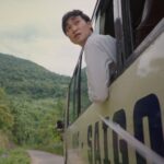 A young Vietnamese man leans out the window of a bus as it passes along a mountain road. He is wearing a white shirt and is looking back at something behind the bus. The bus is beige with the word Saigon in black block letters.