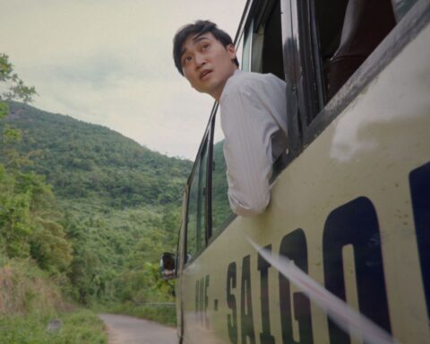 A young Vietnamese man leans out the window of a bus as it passes along a mountain road. He is wearing a white shirt and is looking back at something behind the bus. The bus is beige with the word Saigon in black block letters.