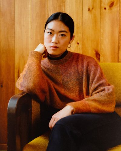 Carol Nguyen is a young Vietnamese-Canadian filmmaker. She is pictured in a portrait, sitting on a couch with her legs crossed and her right arm propped up to her chin. She is wearing a brown shirt and black pants. The wall behind her has wood panelling.