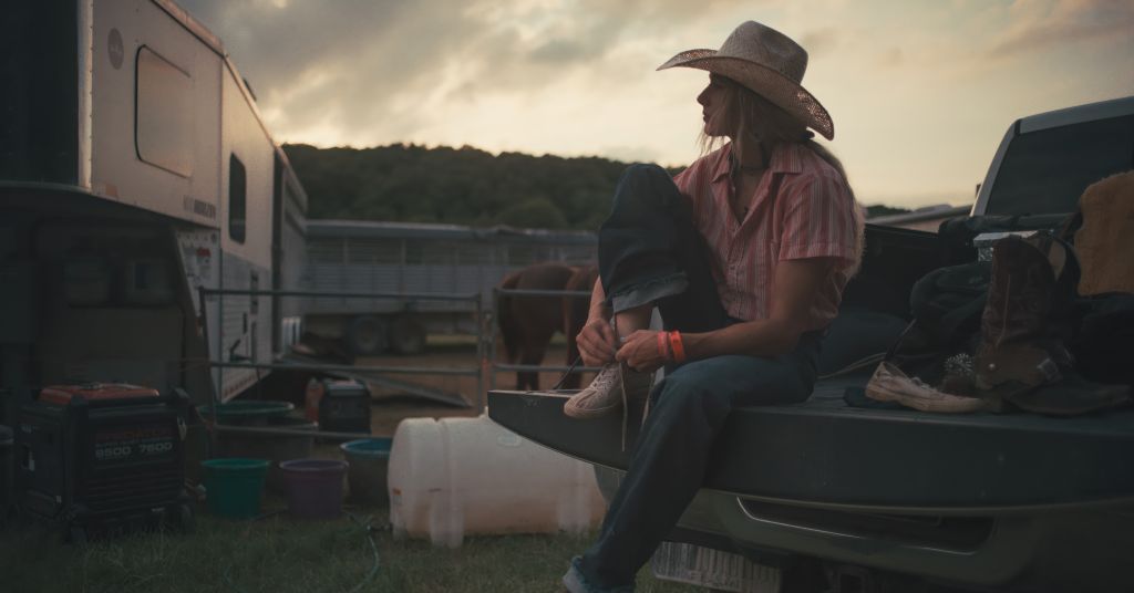 A cowgirl wearing a pink collared shirt and white cowboy hat sits on a fence and looks contemplatively at a cloudy sky.