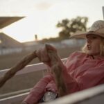 A cowgirl wearing a pink collared shirt and white cowboy hat sits atop a horse.