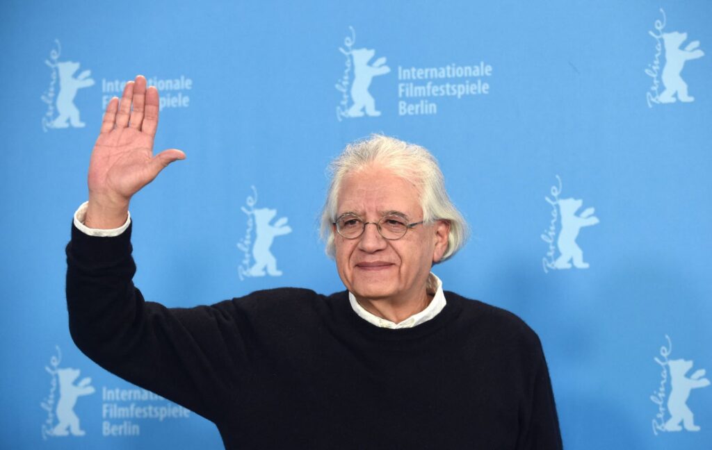 Filmmaker Patricio Guzman is a Chilean filmmaker. He is pictured at the Berlin Film Festival wearing a black sweater overtop a white collared shirt. He has grey hair and glasses. He is waving to someone and smiling. There is a blue media wall behind him.