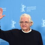 Filmmaker Patricio Guzman is a Chilean filmmaker. He is pictured at the Berlin Film Festival wearing a black sweater overtop a white collared shirt. He has grey hair and glasses. He is waving to someone and smiling. There is a blue media wall behind him.