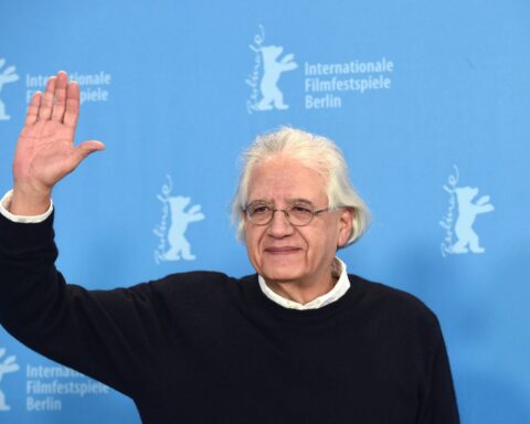 Filmmaker Patricio Guzman is a Chilean filmmaker. He is pictured at the Berlin Film Festival wearing a black sweater overtop a white collared shirt. He has grey hair and glasses. He is waving to someone and smiling. There is a blue media wall behind him.