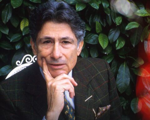 Academic Edward Said looks directly into the camera, posing with this chin on his hand to express thought. He is wearing a suit and there are green leaves behind him. The portrait is bordered by strips of black and white photographs.