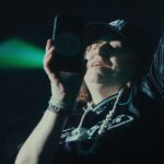 A white woman wearing a black t-shirt and black baseball cap, with chains around her neck, rocks out to a concert in a stadium. She is recording a concert with her phone and singing along.