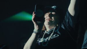 A white woman wearing a black t-shirt and black baseball cap, with chains around her neck, rocks out to a concert in a stadium. She is recording a concert with her phone and singing along.