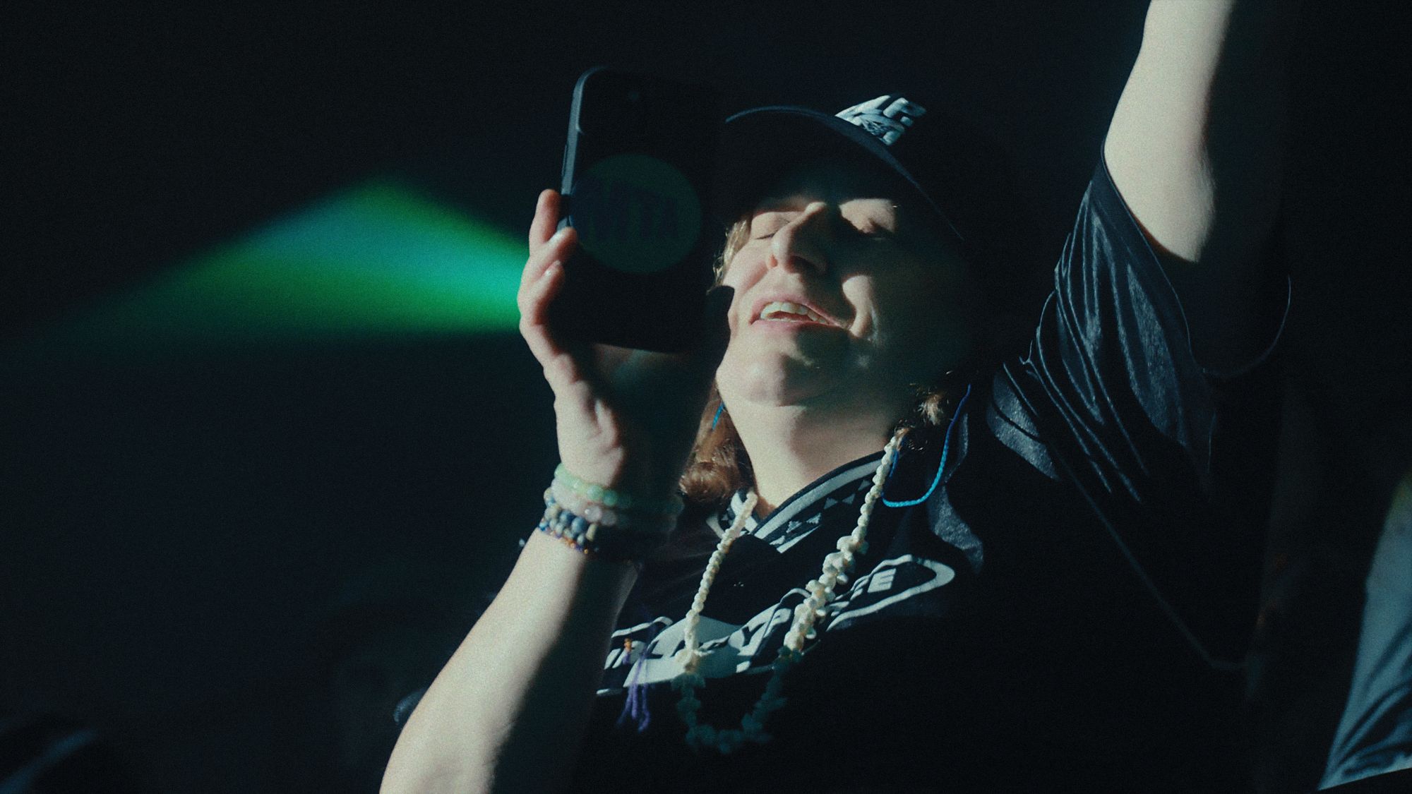 A white woman wearing a black t-shirt and black baseball cap, with chains around her neck, rocks out to a concert in a stadium. She is recording a concert with her phone and singing along.