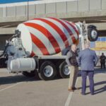 Two men in suits stand at a distance. Behind them is a cement mixer patterned in the style of the American flag. They are standing on a road and a concrete overpass is behind the truck.