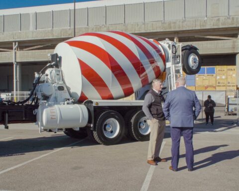 Two men in suits stand at a distance. Behind them is a cement mixer patterned in the style of the American flag. They are standing on a road and a concrete overpass is behind the truck.