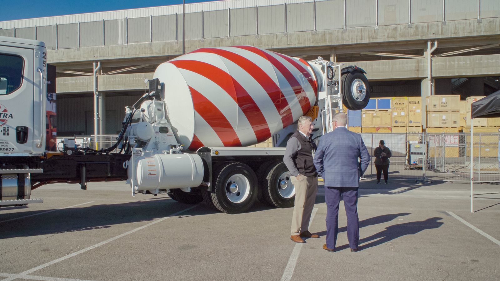 Two men in suits stand at a distance. Behind them is a cement mixer patterned in the style of the American flag. They are standing on a road and a concrete overpass is behind the truck.