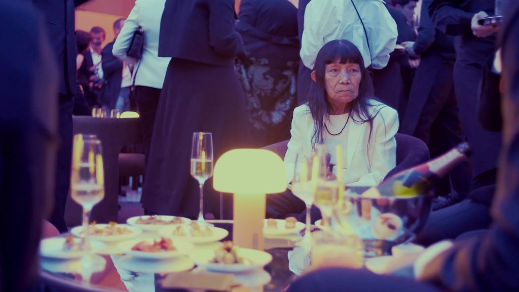 76 year-old Indigenous artist Sara Flores sits along in a busy party. She is wearing a white sleeved dress and there are lights and champagne flutes around her.