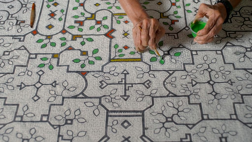 An overhead image of shows artist Sara Flores' hands as she draws Kené, an pattern design of geometric lines in black, green, and red atop a white canvas.