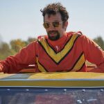 A man wearing an orange jumpsuit, with a yellow V atop the chest and shoulders, stands up through a car's sun roof. He is wearing sunglasses and has his arms spread over the roof of the car.
