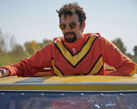 A man wearing an orange jumpsuit, with a yellow V atop the chest and shoulders, stands up through a car's sun roof. He is wearing sunglasses and has his arms spread over the roof of the car.