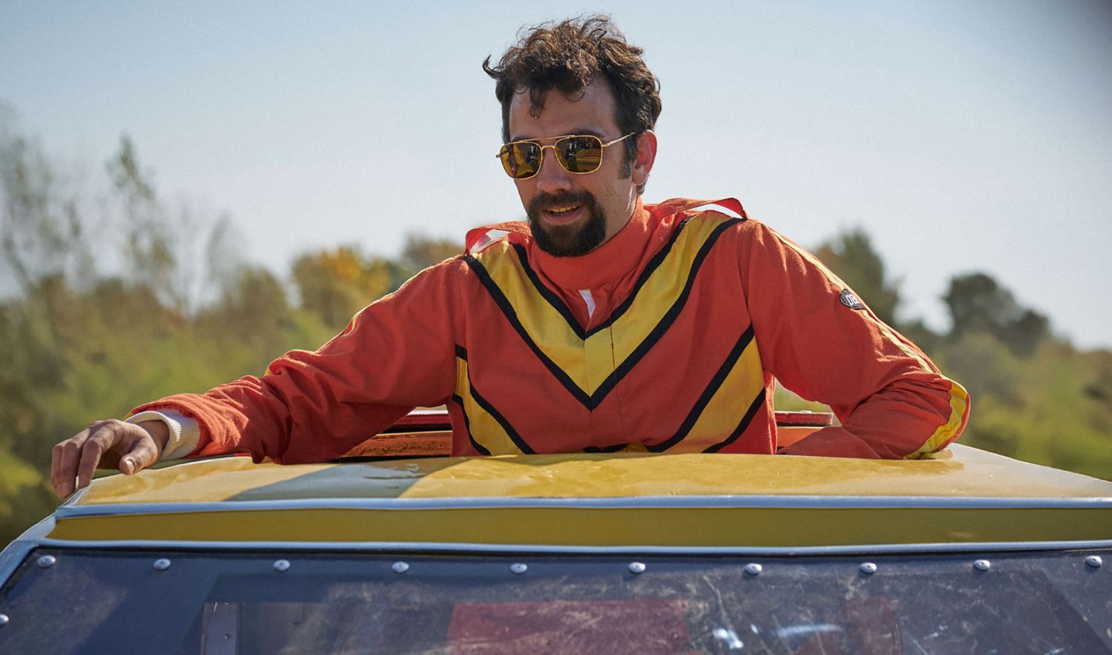A man wearing an orange jumpsuit, with a yellow V atop the chest and shoulders, stands up through a car's sun roof. He is wearing sunglasses and has his arms spread over the roof of the car.