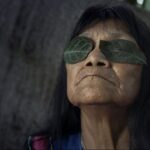 Indigenous artist Sara Flores leans back against a tree with leaves over her eyes. She is pictured in close up. She is a 76-year-old woman with black hair.