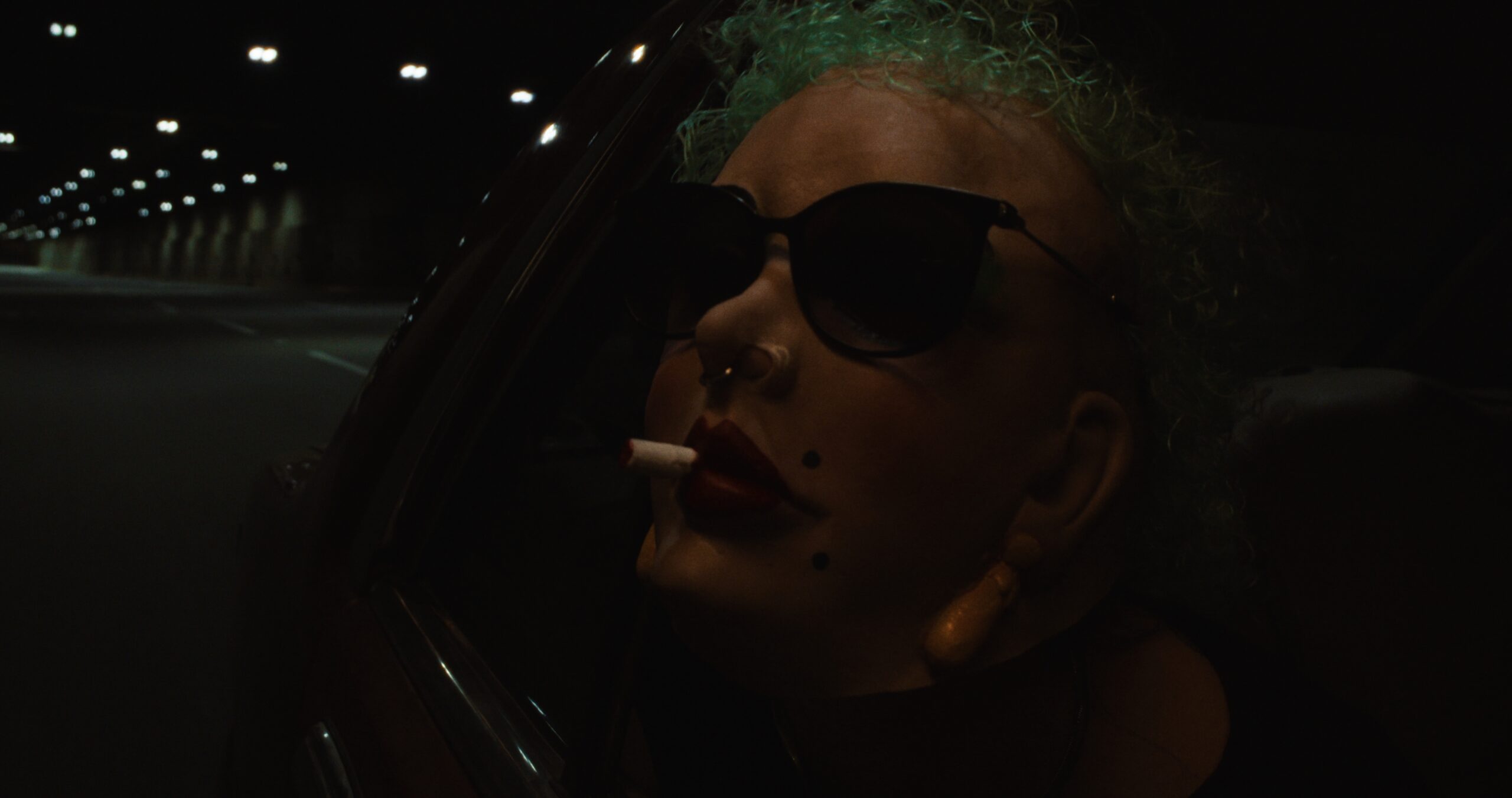 A very dark image offers the obscure sight of a personal wearing a mask with a cigarette dangling from painted lips.