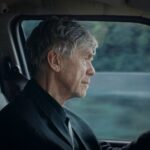 An elderly white man is pictured sitting in the driver's seat of a car. He has short grey hair and is pictured from the point of view of the front passenger seat.