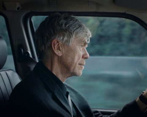 An elderly white man is pictured sitting in the driver's seat of a car. He has short grey hair and is pictured from the point of view of the front passenger seat.