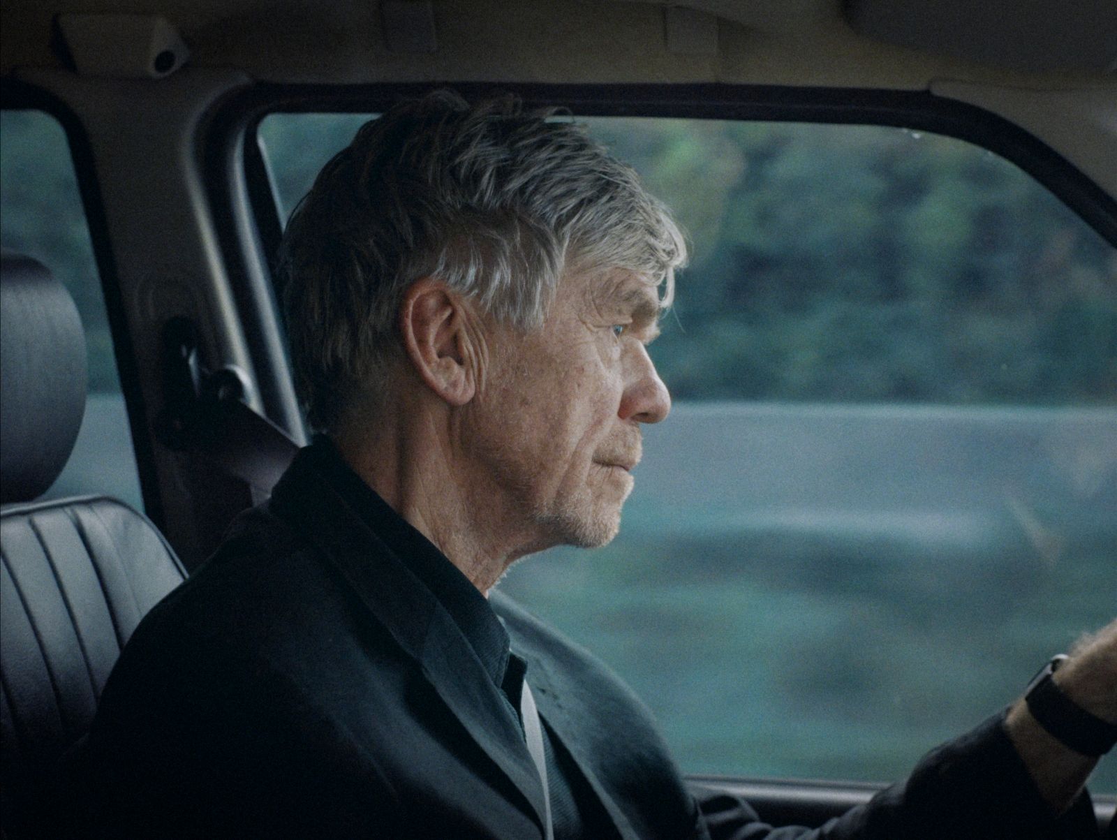 An elderly white man is pictured sitting in the driver's seat of a car. He has short grey hair and is pictured from the point of view of the front passenger seat.