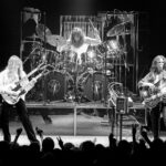 A black and white photo shows the rock band Rush performing on stage.