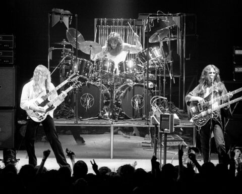 A black and white photo shows the rock band Rush performing on stage.