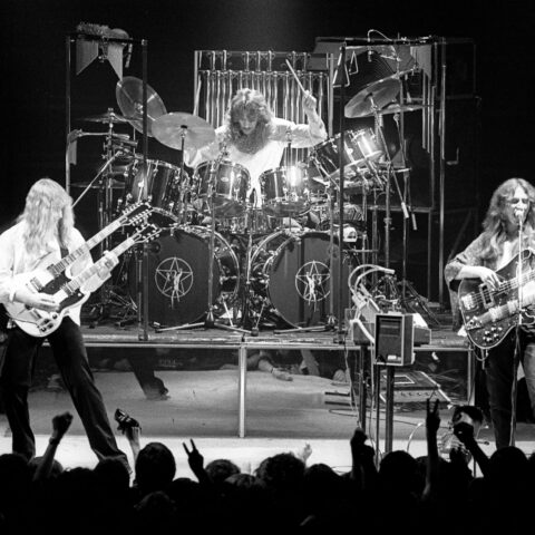 A black and white photo shows the rock band Rush performing on stage.