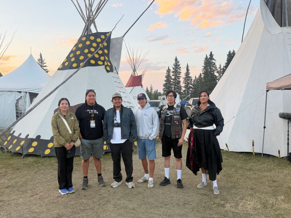 A team of six Indigenous filmmakers stands together in front of several white teepees. The sky behind them is blue with some clouds tinted pink by the sunset.