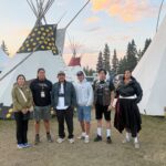 A team of six Indigenous filmmakers stands together in front of several white teepees. The sky behind them is blue with some clouds tinted pink by the sunset.