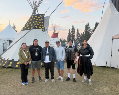 A team of six Indigenous filmmakers stands together in front of several white teepees. The sky behind them is blue with some clouds tinted pink by the sunset.