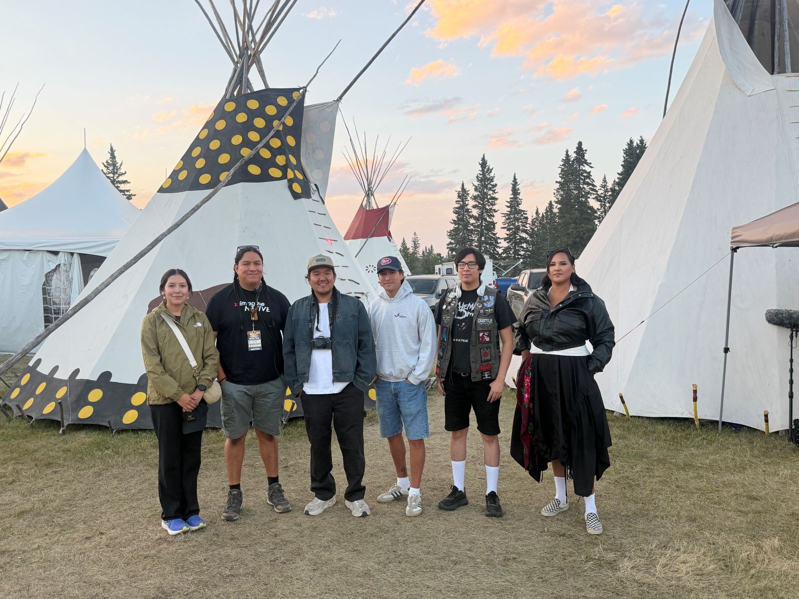 A team of six Indigenous filmmakers stands together in front of several white teepees. The sky behind them is blue with some clouds tinted pink by the sunset.
