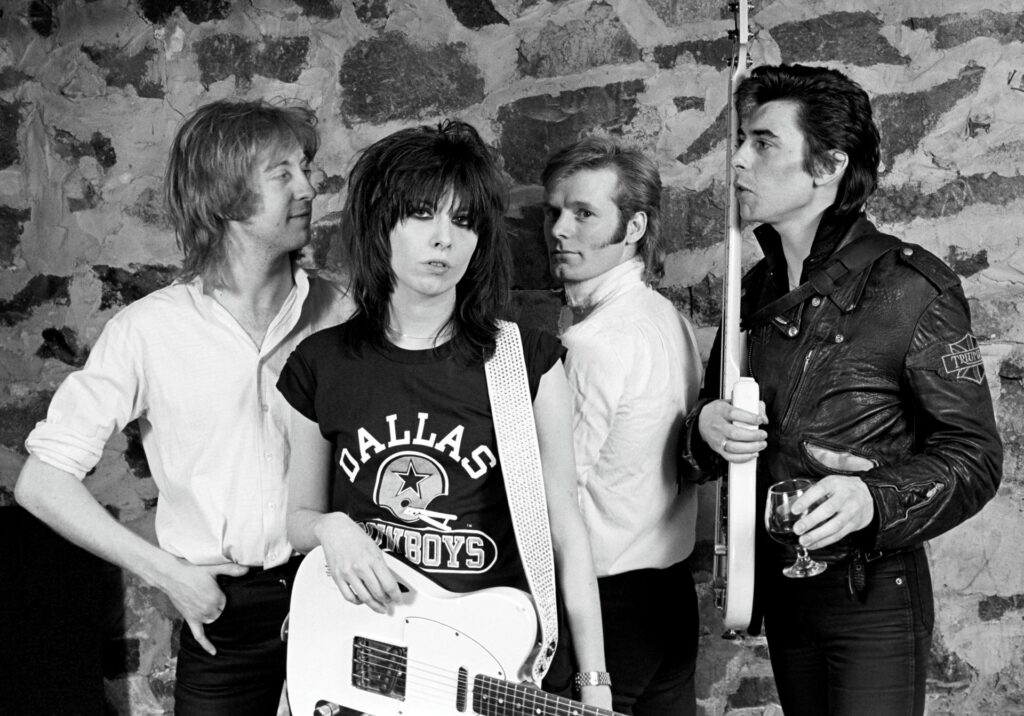 A black and white photo shows the four original members of the Pretenders standing together,holding their guitars and drum sticks. They are in front of a stone wall.