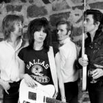 A black and white photo shows the four original members of the Pretenders standing together,holding their guitars and drum sticks. They are in front of a stone wall.