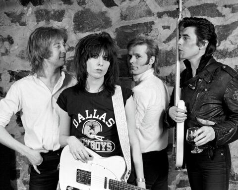 A black and white photo shows the four original members of the Pretenders standing together,holding their guitars and drum sticks. They are in front of a stone wall.