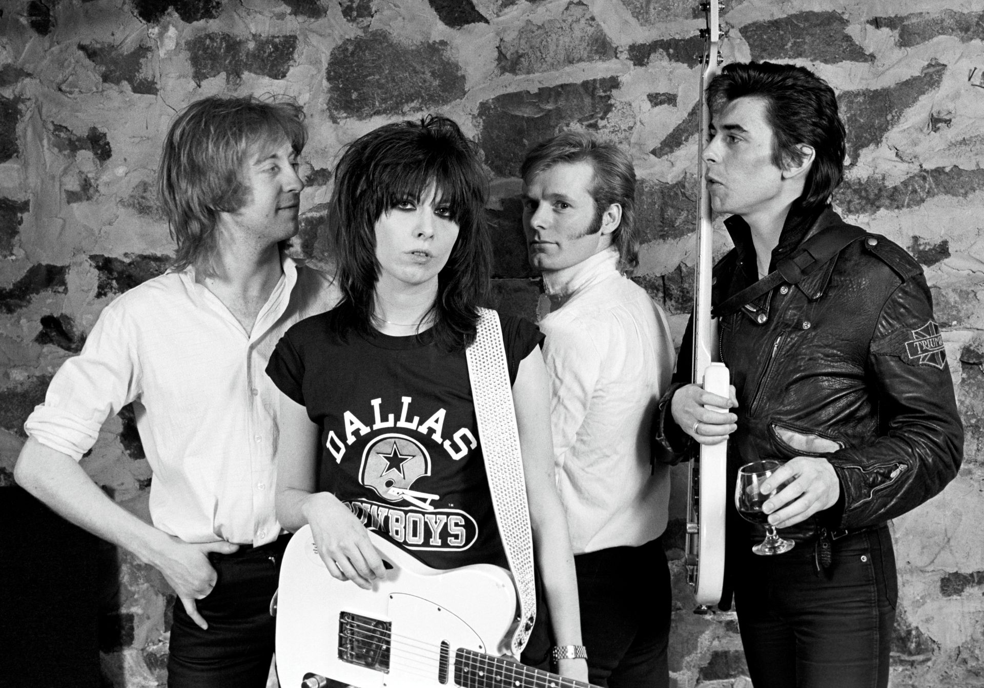 A black and white photo shows the four original members of the Pretenders standing together,holding their guitars and drum sticks. They are in front of a stone wall.