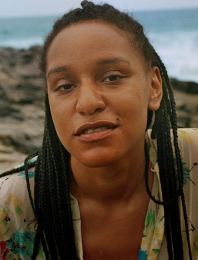 Filmmaker Rebeca Huntt is a young Black woman, pictured in close-up looking into the camera. She has long braided hair and is wearing a light green shirt.