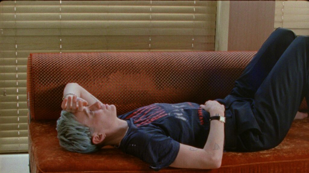 Soccer player Megan Rapinoe lies on her back on a red couch. Rapinoe is a white woman with short cropped purple/grey hair and she is wearing a dark blue t-shirt and black sweat pants.