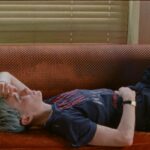 Soccer player Megan Rapinoe lies on her back on a red couch. Rapinoe is a white woman with short cropped purple/grey hair and she is wearing a dark blue t-shirt and black sweat pants.