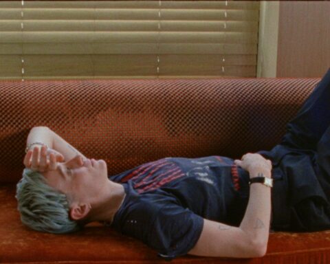 Soccer player Megan Rapinoe lies on her back on a red couch. Rapinoe is a white woman with short cropped purple/grey hair and she is wearing a dark blue t-shirt and black sweat pants.