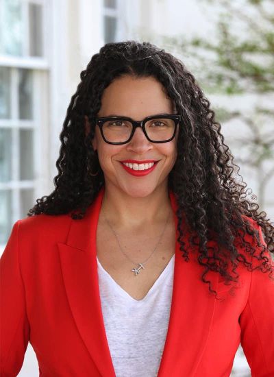 Lacey Schwartz Delgado is a mixed race Black Jewish woman. She has long curly hair and is wearing a red blazer with a white v-neck shirt and black glasses. She is looking at the camera and smiling.